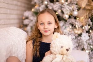 young girl holding teddy bear in front of christmas tree
