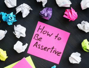 Post it note with How to Be Assertive written on it