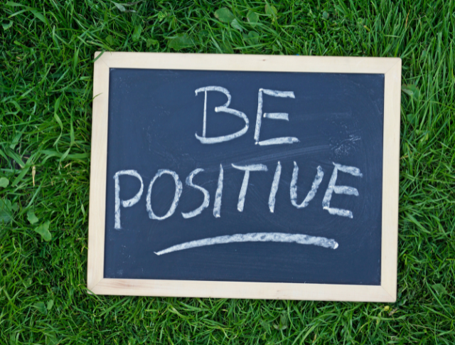 Small blackboard with 'Be Positive' written in white chalk, laid on grass