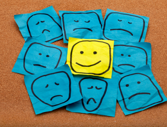 Smiley face drawn on yellow stick note, surrounded by sad faces on blue sticky notes